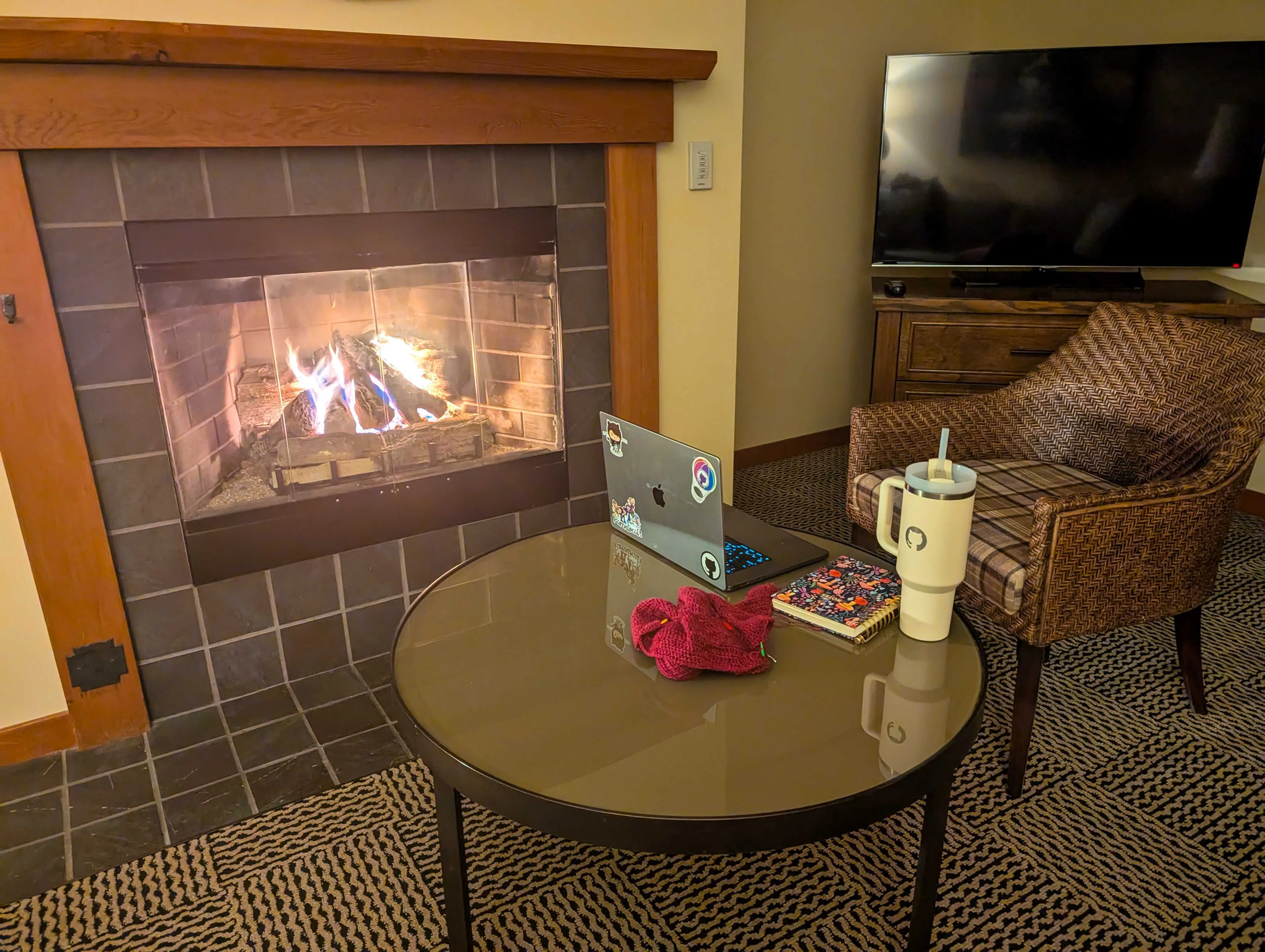 A cozy fireplace with a chair and table in front of it, with a computer open and a crochet project on the table