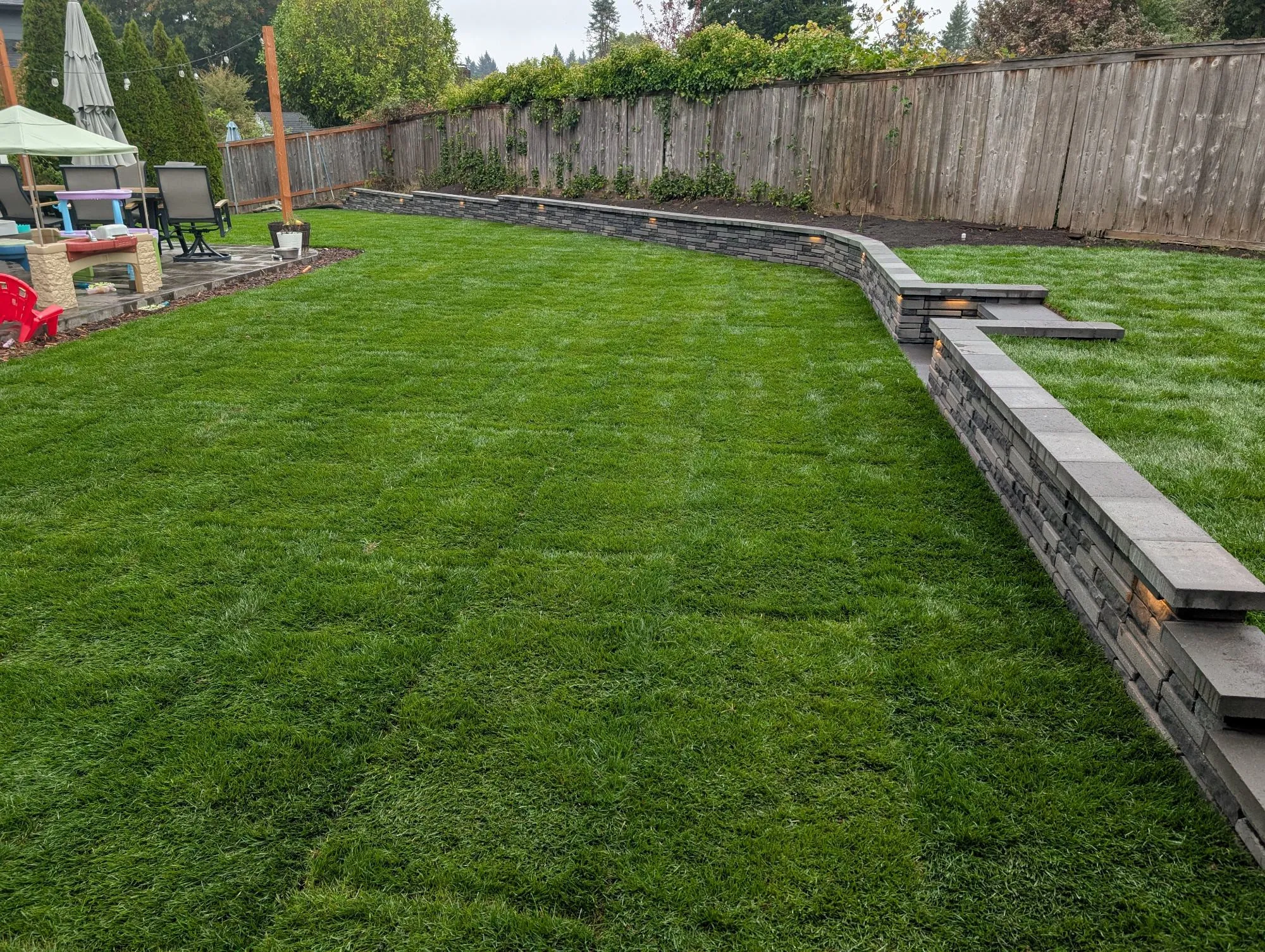 A green backyard with a grey stone retaining wall stretching across it